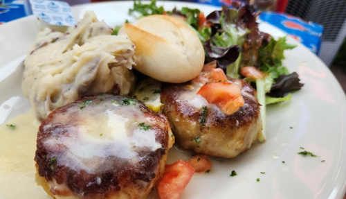 A plate of food featuring two crab cakes, mashed potatoes, a biscuit, and a side salad with mixed greens and tomatoes.