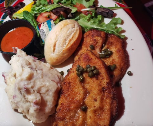A plate featuring two fried fish fillets with capers, mashed potatoes, a roll, and a side salad with dressing.