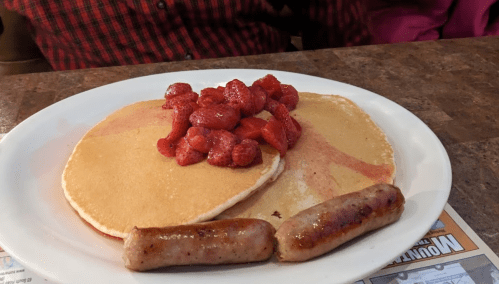A plate with two pancakes topped with raspberries and two sausage links on the side.