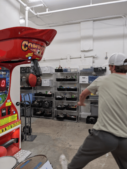 A person swings at a punching bag arcade game in a room with shelves of equipment in the background.