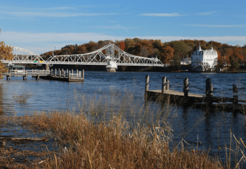 A scenic view of a river with a bridge, autumn foliage, and a white building in the background.