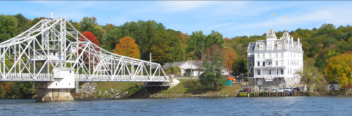 A white bridge spans a river beside a historic white building, surrounded by colorful autumn trees.