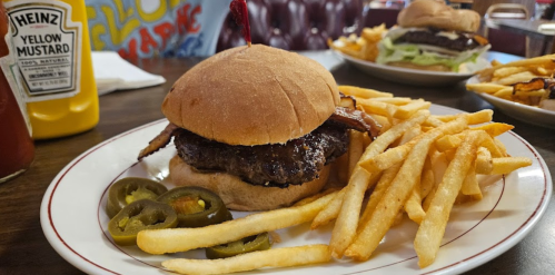 A juicy burger with bacon on a bun, served with fries and jalapeños, alongside mustard and ketchup bottles.