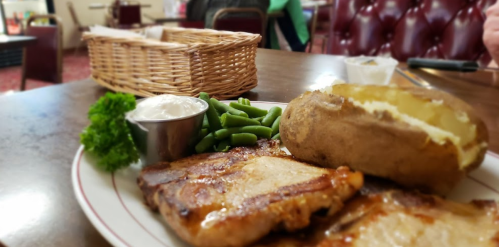 A plate with grilled pork chops, a baked potato, green beans, and a small cup of sour cream, set on a wooden table.