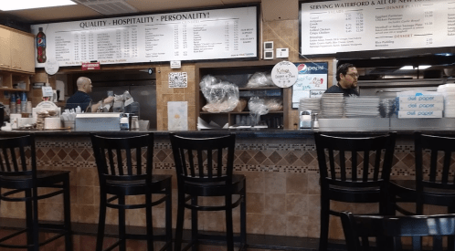 A restaurant counter with two staff members preparing food, menus displayed above, and empty chairs in front.