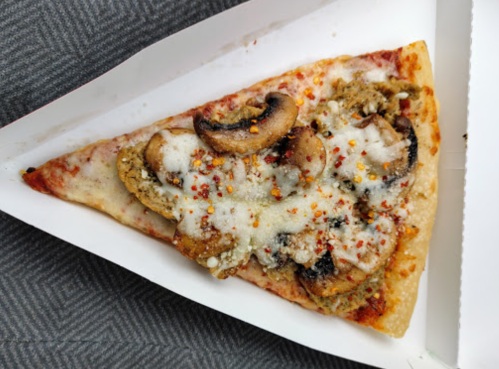 A slice of pizza topped with mushrooms, cheese, and red pepper flakes on a white paper plate.