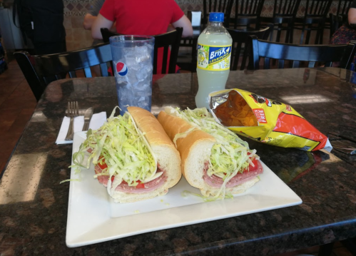 A plate with two large sandwiches filled with lettuce and tomato, a drink, and a bag of chips on a table.