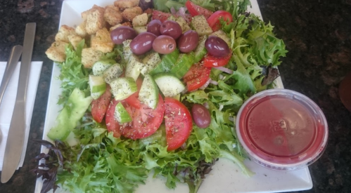 A fresh salad with mixed greens, tomatoes, cucumbers, olives, croutons, and a side of dressing on a white plate.