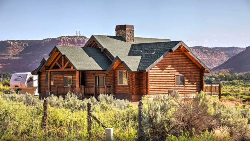 A large log cabin surrounded by greenery, with mountains in the background and a clear blue sky.