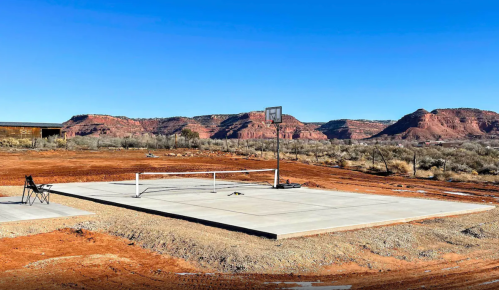 A concrete basketball court in a desert landscape, surrounded by red rock formations and clear blue skies.