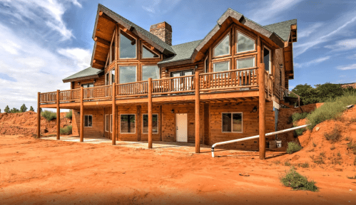 A large wooden house with a balcony, set against a backdrop of red sand and blue sky.