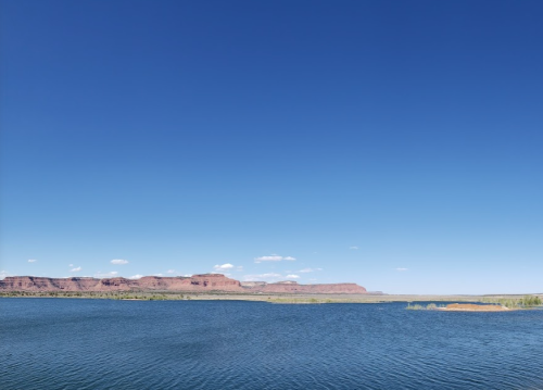 A serene lake with a clear blue sky and distant red rock formations under sunlight.