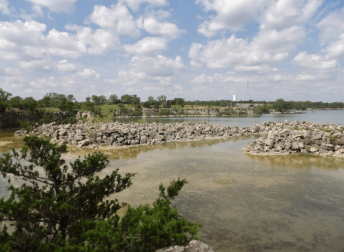 A serene landscape featuring a rocky shoreline, calm water, and a blue sky with fluffy clouds.