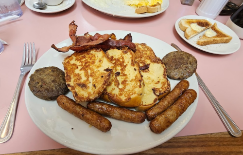 A plate of breakfast featuring French toast, sausage links, sausage patties, and crispy bacon on a pink table.