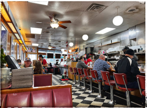 A bustling diner interior with red booths, checkered floors, and patrons enjoying meals at the counter and tables.