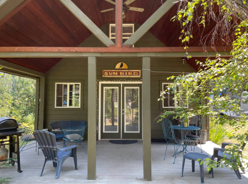 A cozy cabin with a sign reading "Sun Bird," featuring outdoor seating and a grill under a wooden roof.
