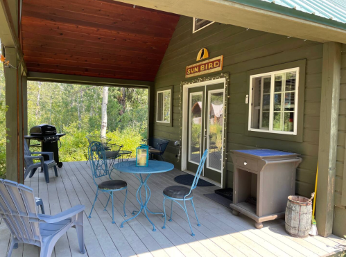 A cozy porch with blue chairs and a table, a grill, and a sign reading "SUN BIRD," surrounded by greenery.