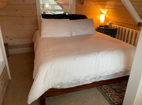Cozy bedroom with a wooden bed, white bedding, and a bedside lamp in a rustic, wooden-paneled room.