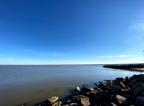 A calm lake under a clear blue sky, with rocky shoreline and distant horizon.