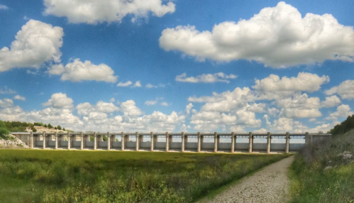 A wide view of a dam with tall concrete pillars under a blue sky filled with fluffy clouds, beside a grassy path.