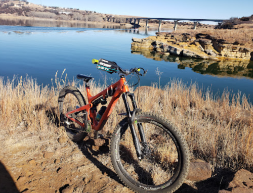 A mountain bike rests on rocky terrain by a calm river, with a bridge visible in the background.