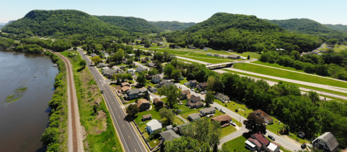 Aerial view of a small town by a river, surrounded by green hills and fields, with a highway running through.