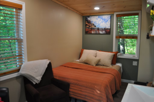 Cozy bedroom with a bed, orange bedding, a chair, and large windows overlooking greenery.