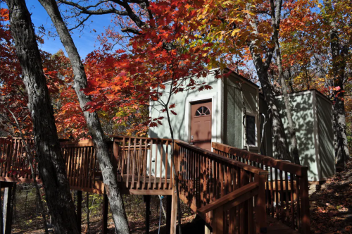 A small cabin surrounded by vibrant autumn foliage and a wooden deck leading to the entrance.