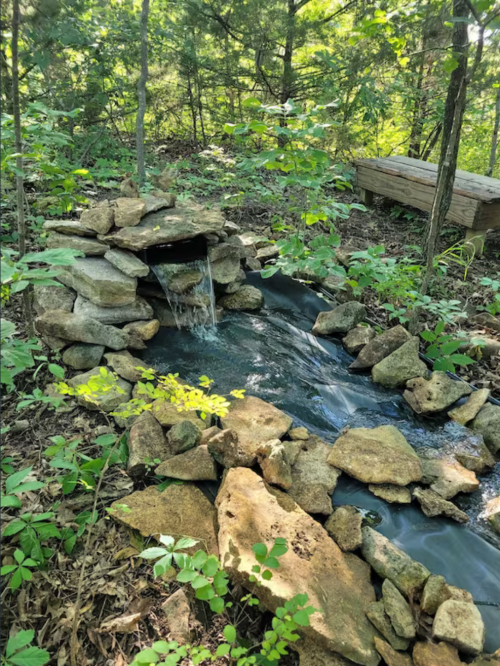 A serene stone water feature flows through a wooded area, surrounded by greenery and natural rocks.