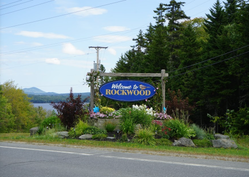 A welcome sign for Rockwood, surrounded by greenery and flowers, with a scenic view in the background.
