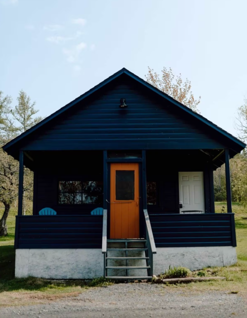 A blue wooden house with an orange door and two chairs on the porch, surrounded by greenery and a clear sky.