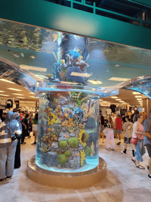 A large cylindrical aquarium filled with colorful coral and fish, surrounded by shoppers in a retail space.