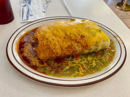 A large burrito topped with melted cheese, served on a plate with green sauce and diced vegetables.