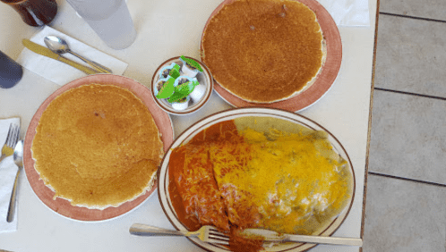 A table with two pancakes and a large plate of enchiladas topped with cheese and sauce, alongside a small bowl of toppings.