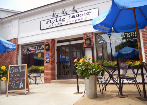 Exterior of Flying Saucer Pizza Company with blue umbrellas, flowers, and a sign that says "We Deliver!"