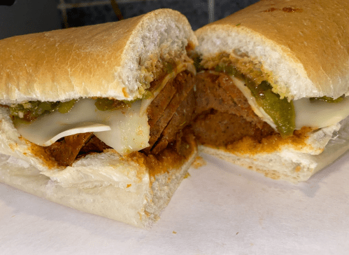A close-up of a sandwich with layers of meat, melted cheese, and green sauce, served in a soft bread roll.
