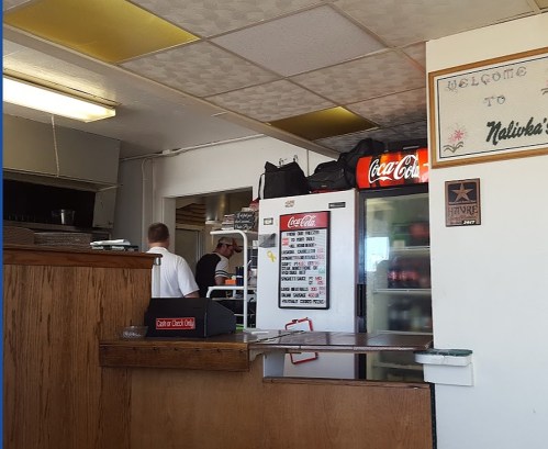 Interior of a diner with a wooden counter, a Coca-Cola fridge, and staff preparing food in the kitchen area.