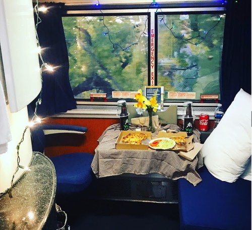 Cozy train interior with a table set for a meal, featuring pizza, drinks, and flowers, with twinkling lights.