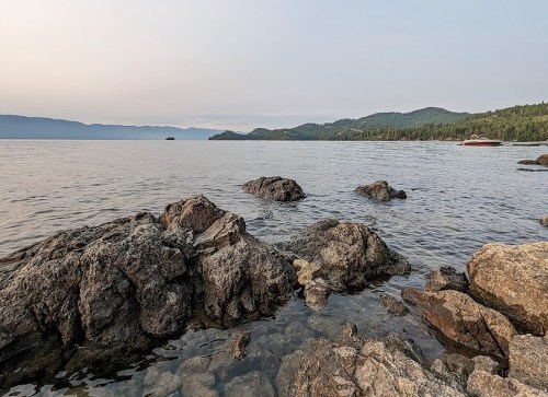 A serene lakeside view with rocky shorelines, calm water, and distant mountains under a hazy sky.
