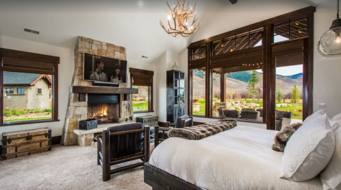 Cozy bedroom with a stone fireplace, large windows, and mountain views, featuring a plush bed and rustic decor.
