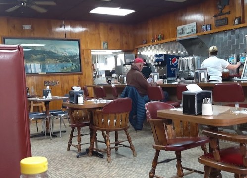A cozy diner interior with wooden walls, tables, and a counter. A customer sits at the counter while a staff member works.