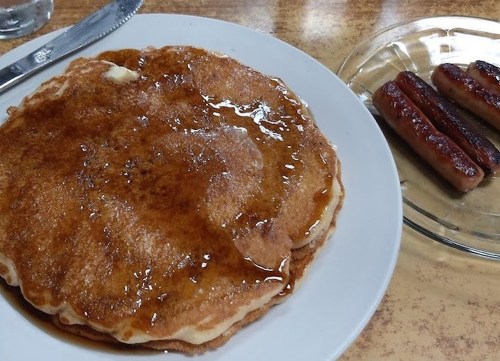 A plate of fluffy pancakes drizzled with syrup, alongside a dish of three cooked sausages.