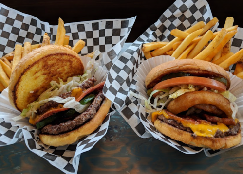 Two burgers with toppings on checkered paper, served with a side of golden fries.