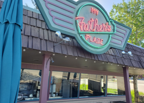 Neon sign for "My Father's Place" on a building with a wooden exterior and outdoor seating area.