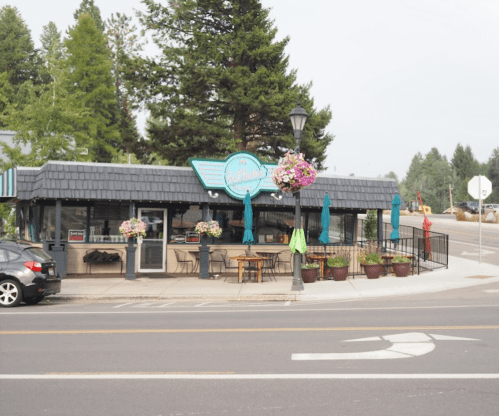 A cozy restaurant with outdoor seating, surrounded by trees and flowers, located at a street corner.