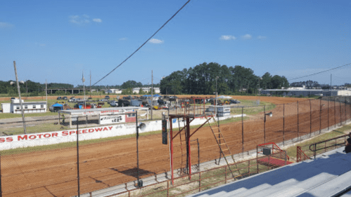 A view of a dirt racetrack with grandstands, pit area, and blue sky in the background.