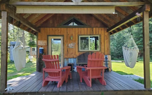 A cozy wooden cabin with two red chairs on a porch, surrounded by greenery and a hammock hanging nearby.