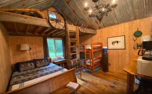 Cozy cabin interior with wooden walls, bunk beds, a double bed, and rustic decor, featuring a chandelier and kitchenette.