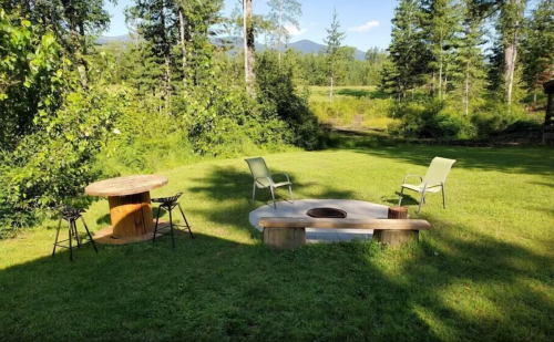 A serene outdoor space with a fire pit, wooden table, and chairs surrounded by trees and mountains in the background.