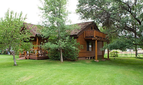 A charming log cabin surrounded by green grass and trees, featuring a balcony and a welcoming porch.